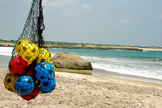 Colorful balls hang on a beachside net with waves, sand, and ocean in the background.