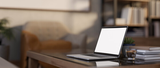 Blank screen laptop aside coffee cup and books on glass top wooden table in a blurred living room.
