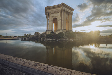 View of architectural marvel stands majestically on a small island, its reflection shimmering in the still water under a dramatic sky, Montpellier, Occitanie, France.