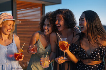 Four cheerful young women laughing and drinking cocktails at sunset party