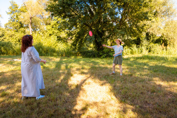 Fototapeta premium Happy Caucasian woman and her daughter play frisbee on the grass during holiday in a public park. Family activity, summer mood and bonding through motion.