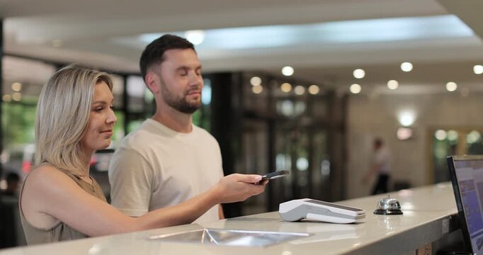 Woman makes nfc payment with mobile phone at pos terminal, paying for room at hotel. Person at reception desk and doing check.