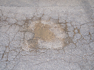 Close up abstract textured background of cracked asphalt with a central worn patch of granular dirt and small stones revealing underlying surface imperfections and wear patterns 