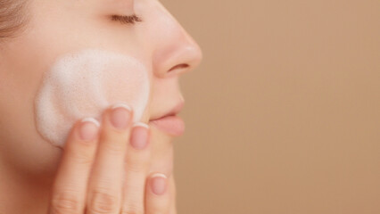 Close-up of a beautiful female face. Woman applying cleansing foam gently washing face ads. Bubbles. Beauty daily routine. Commercial shoot. Skincare treatment. Selfcare. Copy space. Beige Background