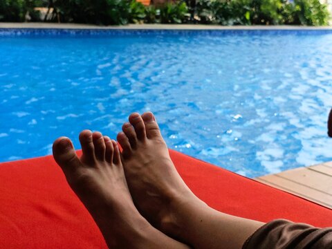Relaxing Poolside Bare Feet on Red Lounger with Sparkling Blue Water.