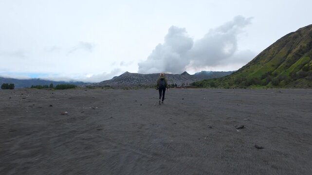 Woman walks alone across black volcanic sand toward lush Mount Batok and smoking Mount Bromo in Indonesia. Scenic travel moment filled with solitude, nature, and adventure.