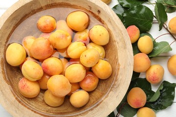 Fresh ripe apricots in bowl with water and green leaves on white wooden table, flat lay