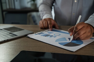 Close-up of online entrepreneur analyzing data charts on paper with stylus pen next to laptop, representing digital business strategy, remote work, and modern financial planning.