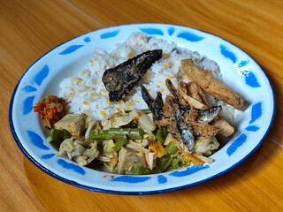 A portion of typical Indonesian mixed rice served on a classic blue and white patterned enamel plate, consisting of corn rice, a piece of dried fish, fried tempeh, stir-fry and chili sauce. Delicious 