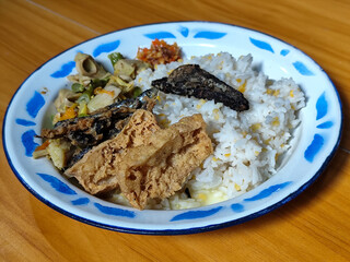A portion of typical Indonesian mixed rice served on a classic blue and white patterned enamel plate, consisting of corn rice, a piece of dried fish, fried tempeh, stir-fry and chili sauce. Delicious 
