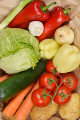 A close-up, overhead view displays a rich variety of fresh, raw vegetables including red and yellow bell peppers, lettuce, zucchini, carrots, tomatoes, onion, garlic, and potatoes, densely packed with
