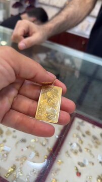 Close-up video of a hand holding a rectangular gold pendant featuring a detailed embossed design, displayed inside a jewelry store.