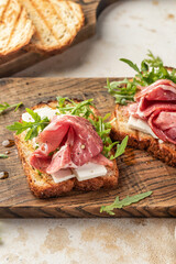Grilled toasts with ham, cheese and arugula on wooden cutting board on light textured background close up. Delicious breakfast or snack