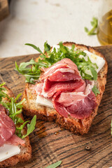 Grilled toasts with ham, cheese and arugula on wooden cutting board on light textured background close up. Delicious breakfast or snack