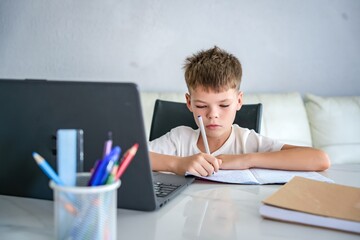 Boy doing homework at home, writing in notebook and studying online on laptop