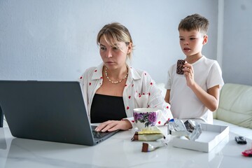 Mother working on laptop while son stands nearby eating chocolate