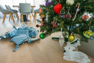 Artificial plastic Christmas tree stands near heap of ripped wrapping paper. A modern festive interior reflecting overconsumption, environmental impact, and unsustainable seasonal traditions. 