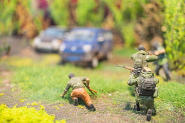 Diorama scene of soldiers advancing towards a village, one looking down at an object, both carrying guns and backpacks Outdoor setting during daylight with muted earthy tones and realistic style Lo