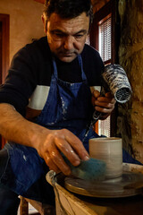 Close-up of male potter working on pottery wheel, shaping ceramic mug with sponge and heat gun