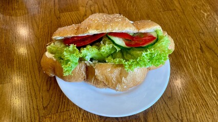 Fresh croissant sandwich with lettuce, tomato, cucumber and mayonnaise on a white plate, served on wooden table, close-up view of healthy vegetarian breakfast or lunch