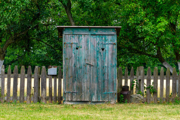 Old wooden outhouse standing in front of a rustic fence, surrounded by green trees in a rural landscape.