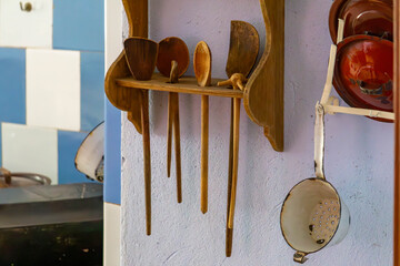 Wooden spoons, ladles, and a strainer hanging on the wall in a vintage kitchen, next to a shelf with enamel dishes and a blue-and-white tiled wall