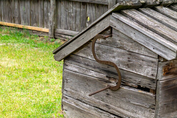 Old wooden well with a manual iron handle for drawing water, set against a wooden fence and grassy yard. Rustic element of a rural homestead.