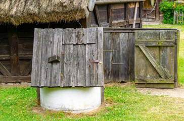 Historic wooden well with a pitched roof and hinged cover, standing on the grass next to a thatched cottage, surrounded by traditional rural wooden buildings.