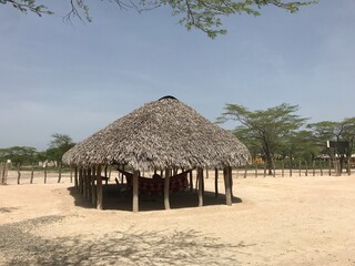 A thatched - roof hut in Riohacha, La Guajira, Colombia. Built with wooden posts, it shelters a colorful Wayuu hammock, set in the arid, tree - dotted tribal land.