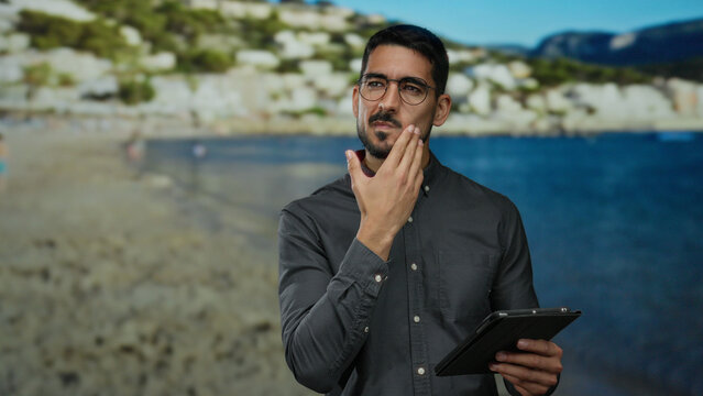 Young man with a thoughtful expression holding a tablet on a beach background with the sea in view.