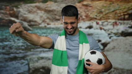 Naklejka premium Young man at seaside beach holding soccer ball wearing green scarf looking enthusiastic and cheerful.