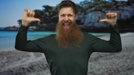 Man with long beard wearing green shirt laughing and pointing thumbs to chest at seaside beach;...