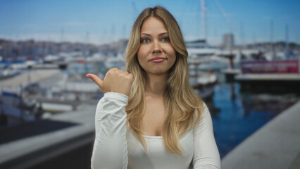 Obraz premium Woman with long blonde hair in white top pointing bare thumb toward docked sailboat at sunny port; indecision hesitation.