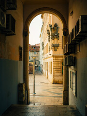 Fototapeta premium Historic stone passage in Republic Square, Split, Croatia, frames a narrow tiled street with ornate facades, leading to Neretvanska ulica under morning light.