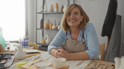 Woman folded hands at ceramics table inside art studio surrounded by clay tools, pottery vases and paintbrushes; contentment.
