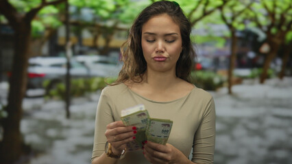 Woman counting argentine pesos with a contemplative expression in a vibrant outdoor park setting, surrounded by lush trees and natural light.