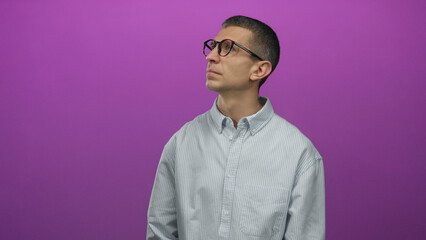 Obraz premium Man in glasses and striped shirt looks up against a purple wall, isolated from the background, showcasing curiosity and individuality in a minimalist environment.