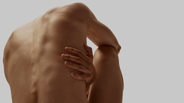 Curved male back with arm wrapped around torso against gray background. Concept of body and emotions, psychosomatic stress, modern masculinity, somatic practice.