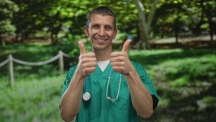 Man doctor smiling outdoors in park wearing green uniform with stethoscope, giving thumbs up, expressing positivity and health awareness in a natural setting.