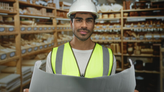 Man holds large blueprint and inspects project plan in studio among wooden shelves filled with wood samples; concentration.