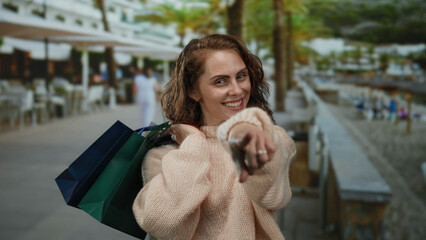 Woman smiling holding shopping bags on a sunny outdoor promenade by the seaside, embodying joy in leisurely beachside shopping.