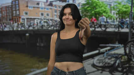 Naklejka premium Young hispanic woman in black crop top and denim jeans gives thumbsdown gesture on a city canal walkway lined with bicycles; disapproval.