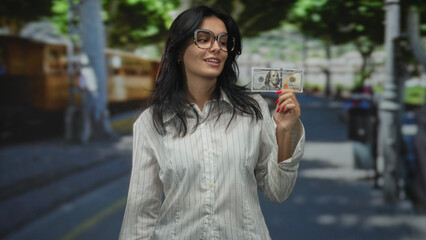 Woman wearing glasses holding hundred dollar bill on street paved sidewalk under green trees; wealth confidence.