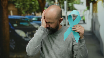 Bald man with beard holding blue cancer ribbon while standing in an urban street, expressing...