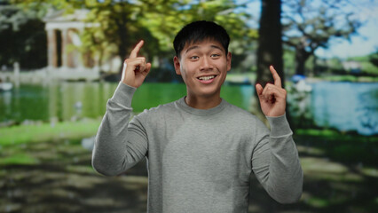 Young man smiling and pointing, standing in a lush outdoor park setting with vibrant greenery and trees, conveying happiness and positivity.