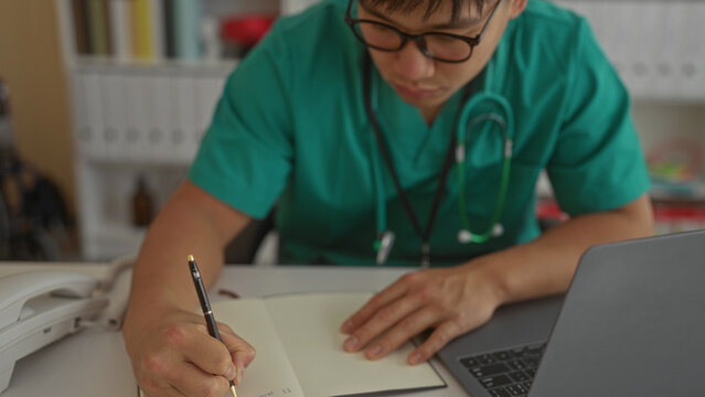 Young doctor man writing notes in clinic room with laptop on desk for professional documentation and medical record maintenance featuring hospital interior and workplace setting