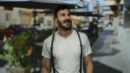 Fototapeta premium Hispanic man with beard smiling outdoors on a lively street, wearing a white shirt and suspenders, surrounded by vibrant urban scenery, conveying joy and warmth in the moment.