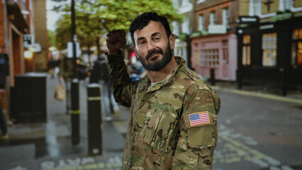 Fototapeta premium Hispanic man in military uniform with beard stands on a vibrant street outdoors, showcasing urban life and american pride with a flag patch on his sleeve.