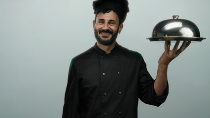 Hispanic chef man with a beard smiling while holding a silver tray against a grey background in a...
