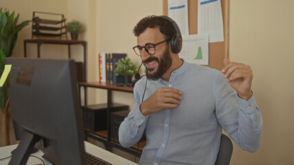 Naklejka premium Hispanic man with beard wearing headset in office, expressing happiness during a video call in a professional environment featuring a computer and office supplies visible.